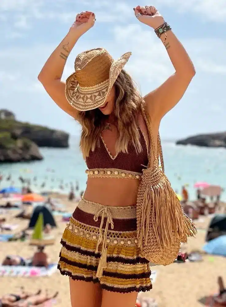Mulher com look boho na praia, usando biquíni e saia de crochê com acessórios, sorrindo e dançando ao ar livre sob o sol.