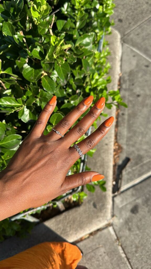 A imagem de uma mão com as unhas pintadas de laranja vibrante, com o sol batendo.