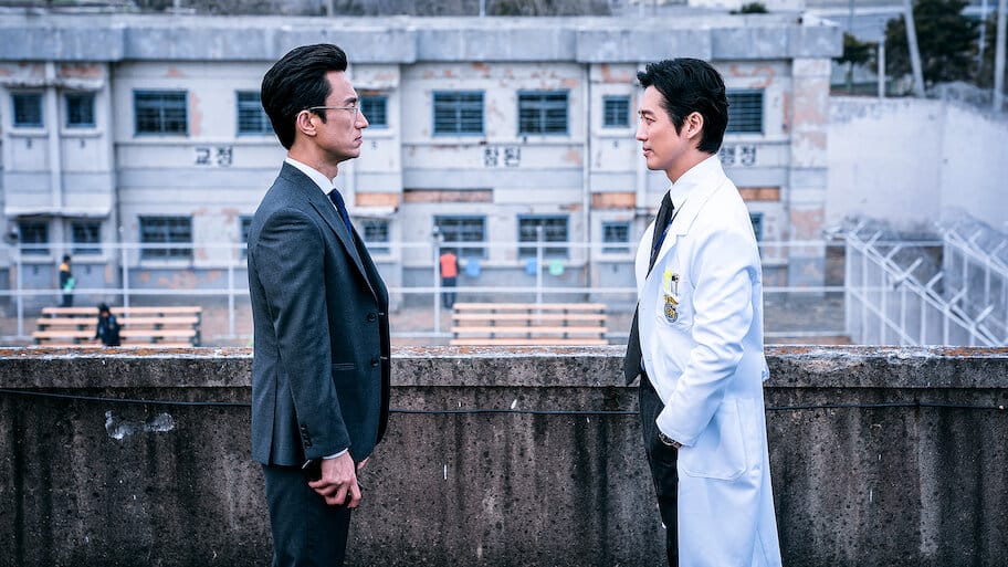Os protagonistas do dorama de suspense 'Doctor Prisoner' se encaram em um telhado com o pátio de uma prisão ao fundo, um vestindo um terno e o outro um jaleco de médico.