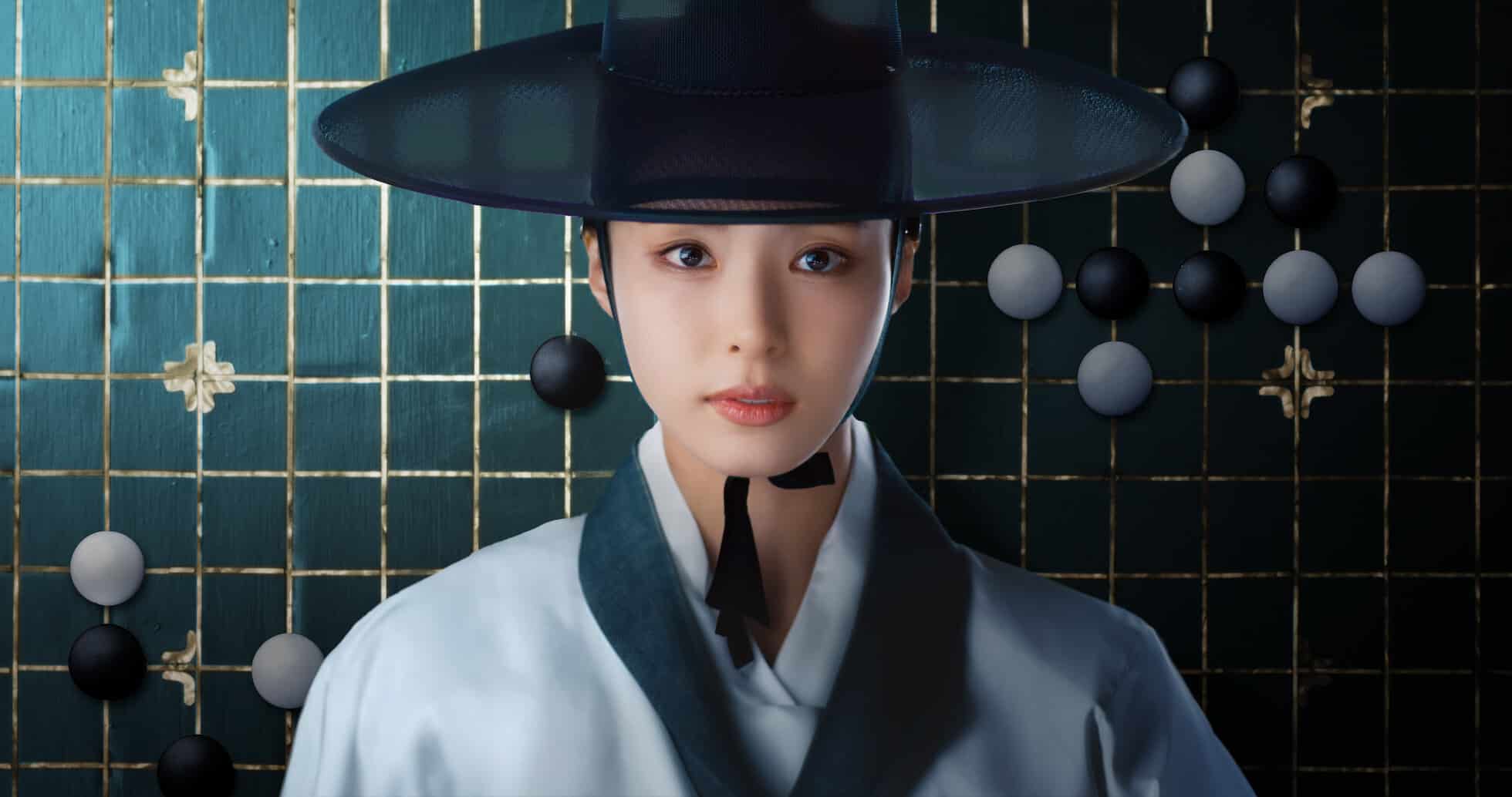 A atriz Shin Se-kyung em trajes masculinos tradicionais, olhando diretamente para a câmera, com um tabuleiro de Go ao fundo, cena da série "Reino da Conquista".