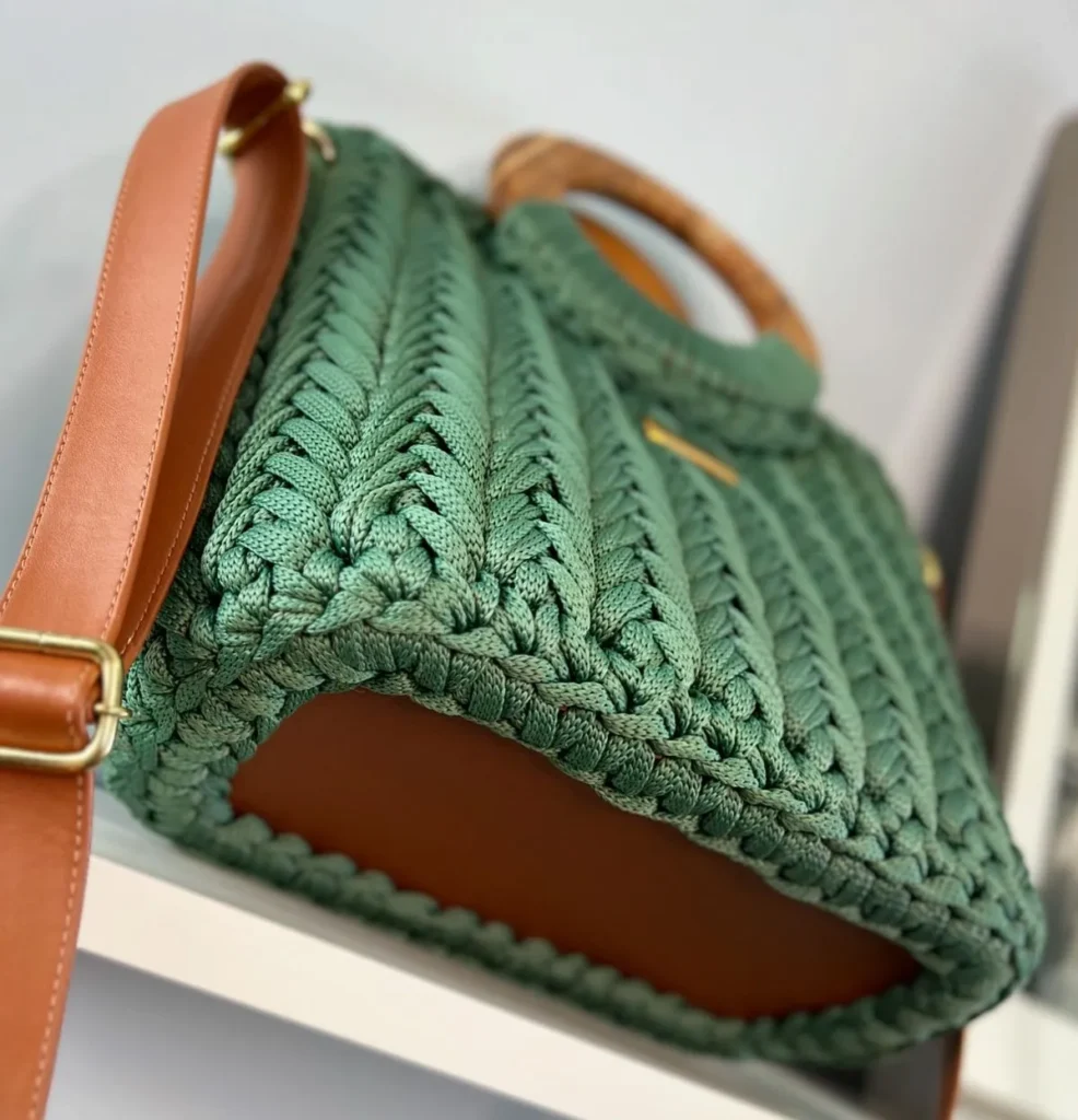 Bolsa de couro com base de couro, alças de madeira e detalhes em fio trançado verde.
