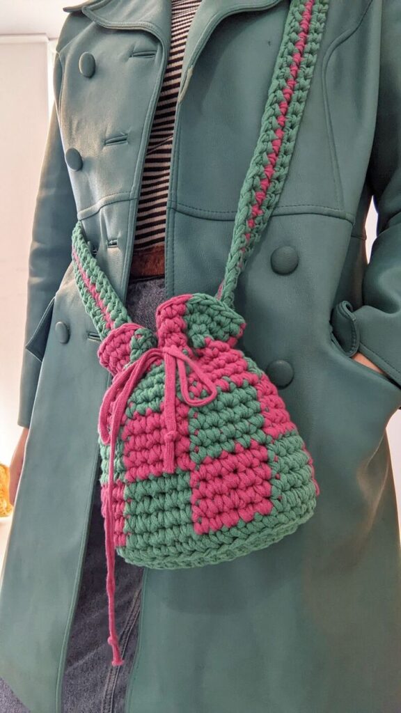 Mulher com um casaco verde-azulado, segurando uma bolsa de crochê estilo 'bucket' em tons de verde e rosa.
