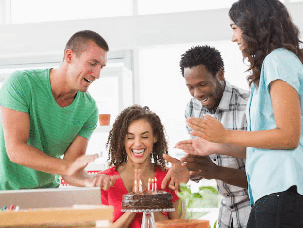 Mulher sorrindo de forma espontânea e feliz enquanto três amigos a surpreendem com um bolo de aniversário, aplaudindo ao redor da mesa.