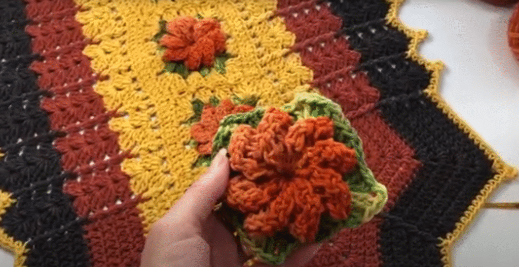 Mão segurando um aplique de crochê sobre um tapete com padrão em degradê de cores quentes.