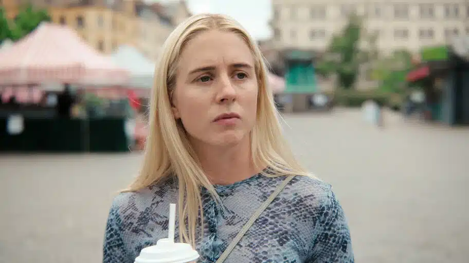 Mulher loira segurando um copo, com expressão pensativa, em uma praça.