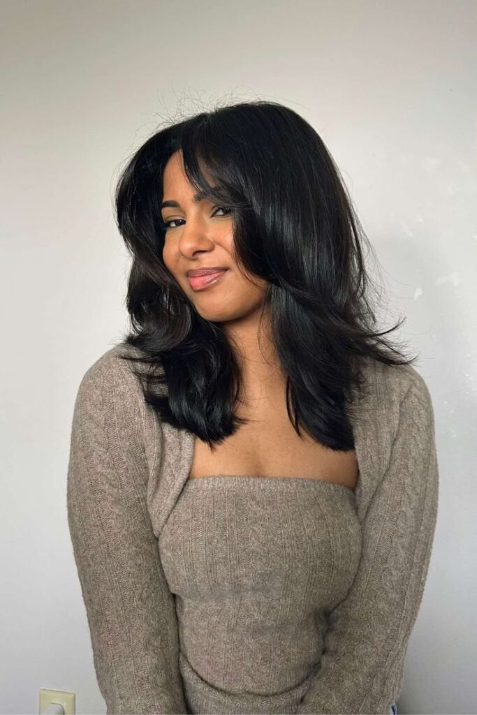 Mulher jovem com cabelos castanhos escuros e o corte de cabelo "butterfly", sorrindo levemente e olhando para a câmera.