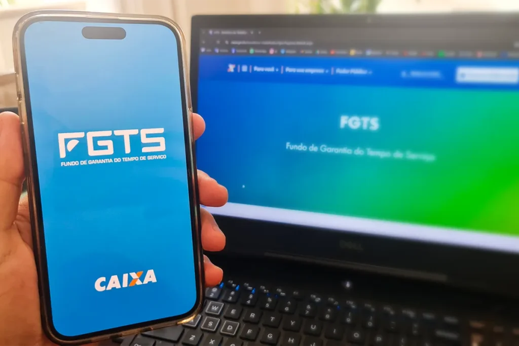 Pagamento FGTS Aplicativo e Dinheiro Uma mão segura um maço de notas de R$ 100 ao lado de um smartphone que exibe o logo do FGTS. A imagem simboliza a regularização financeira de débitos relacionados ao Fundo de Garantia.