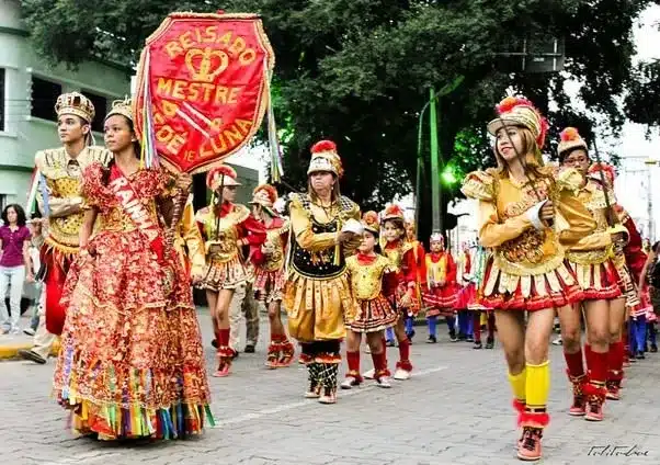 Grupo de pessoas participando da Folia de Reis com trajes coloridos, dançando e carregando um estandarte durante a celebração.