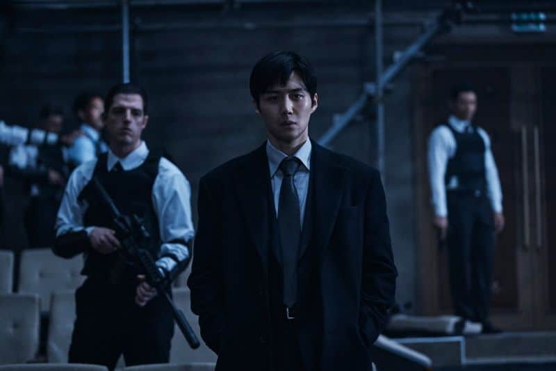 O ator Kim Seon-ho em uma cena do filme de ação "Operação Tyrant", vestido de terno, com seguranças ao seu redor.