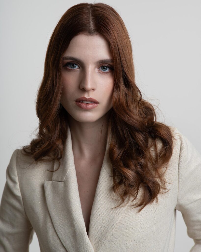 Retrato da atriz Ana Ruggiero, com cabelos ruivos ondulados e olhos azuis, vestindo um blazer claro.