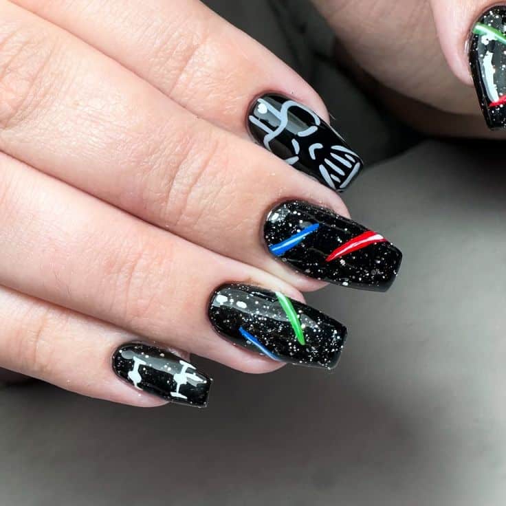 Unhas decoradas com nail art inspirada em Star Wars, incluindo o capacete de Darth Vader, sabres de luz e naves espaciais, em um fundo de esmalte preto com glitter.