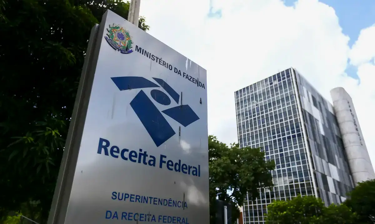 Placa da Receita Federal com logotipo e edifício ao fundo, sede do Ministério da Fazenda.
