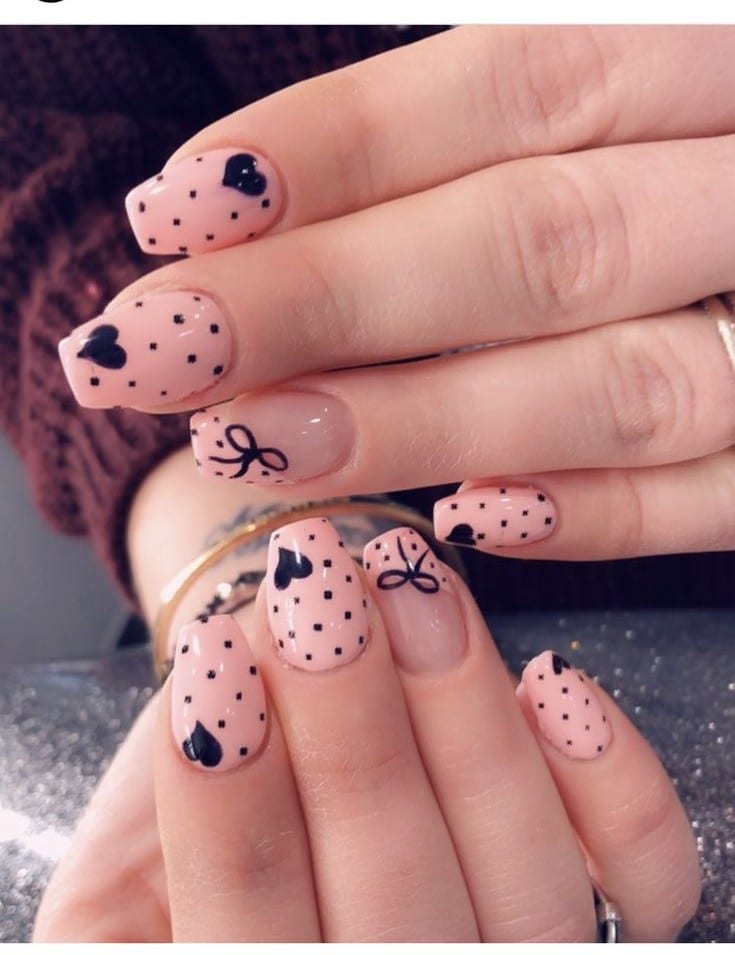 Mãos com unhas decoradas com esmalte rosa claro, bolinhas pretas (poá), corações e laços desenhados, oferecendo um estilo divertido.