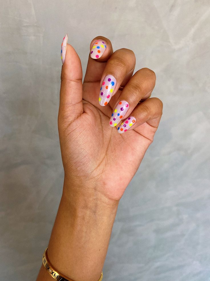 "Mão com unhas curtas decoradas com poás neon em tons de rosa, amarelo e laranja sobre base branca, com um fundo desfocado.
