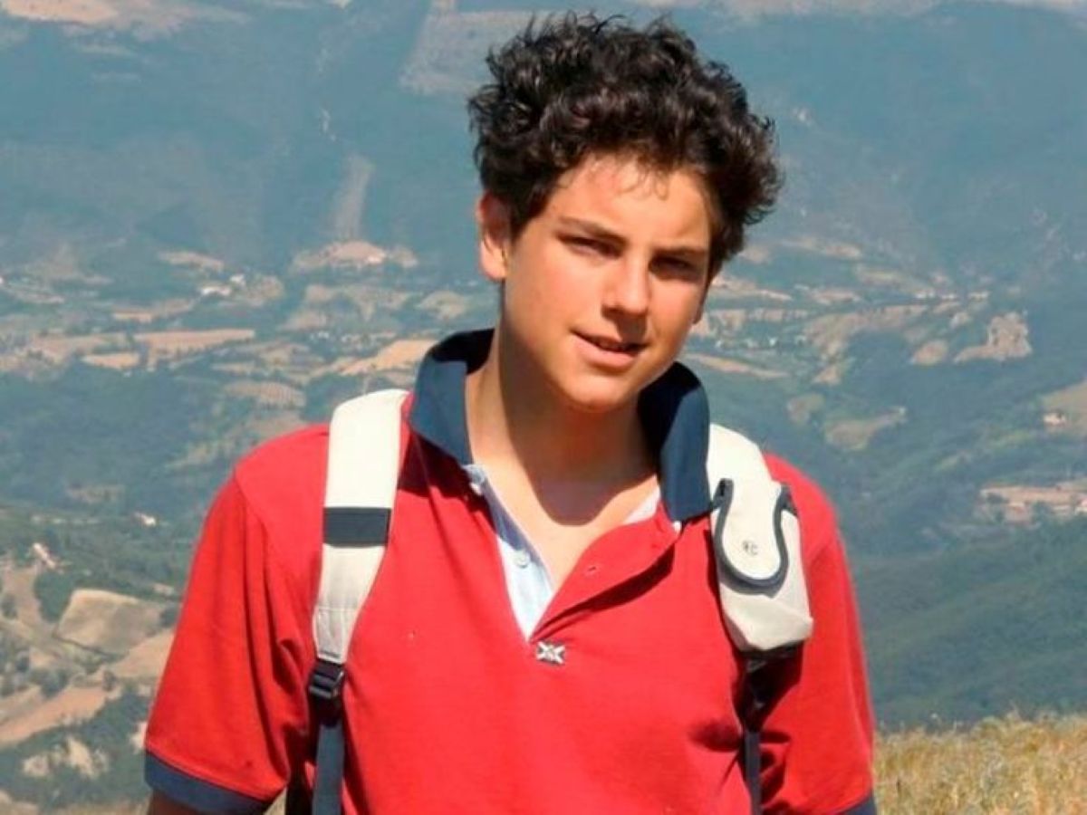 Jovem sorrindo, Carlo Acutis, em um campo verde com montanhas ao fundo, usando uma mochila e uma camisa polo vermelha.