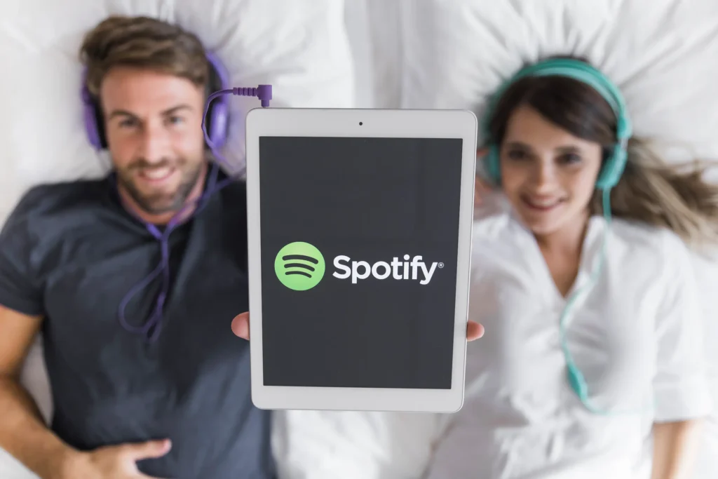 Imagem mostrando um tablet com o logo do Spotify enquanto dois usuários, cada um com fones de ouvido, estão relaxando em uma cama.