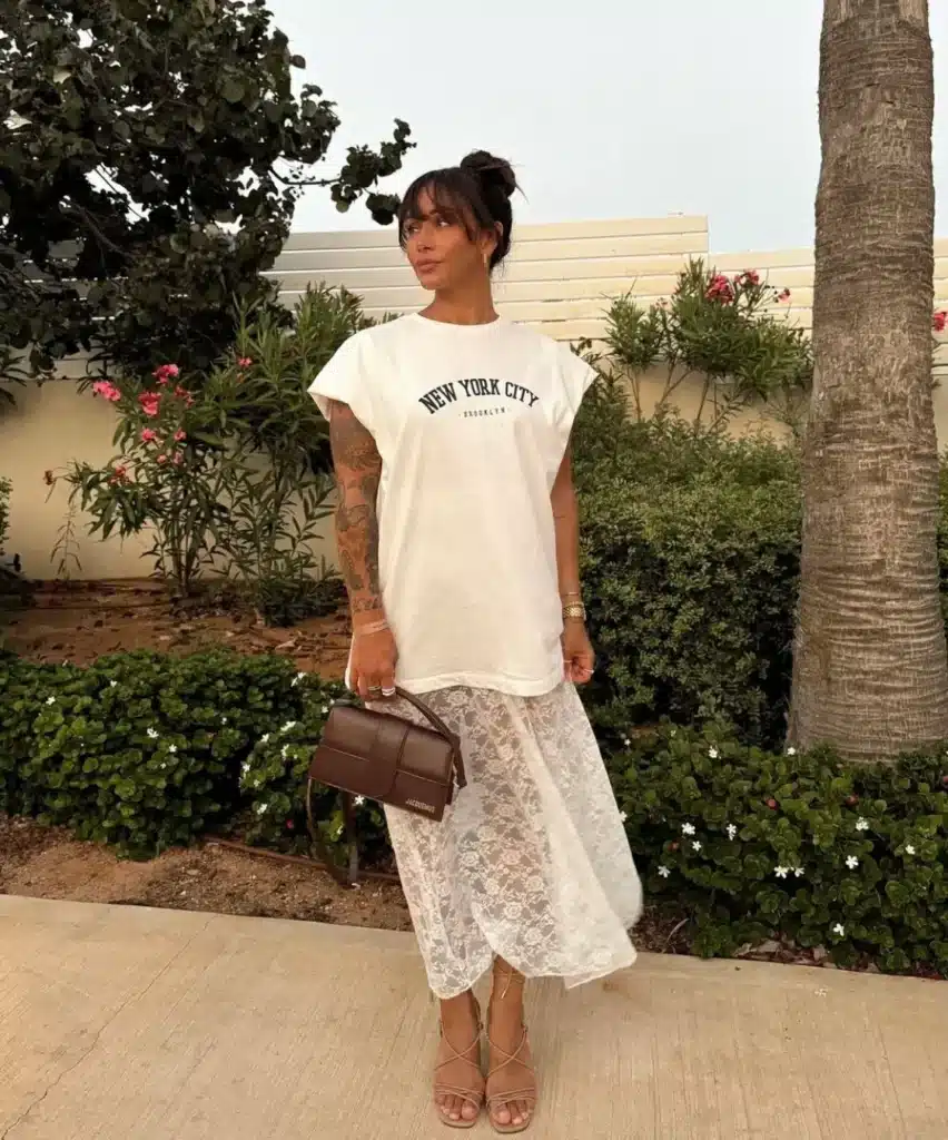 Look urbano com camiseta oversized e saia de renda Mulher usando camiseta oversized branca com estampa 'New York City' e saia de renda transparente branca em ambiente ao ar livre.