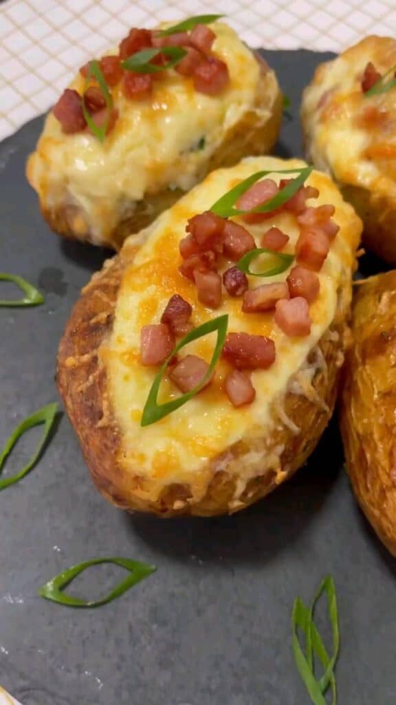 Batata recheada de forno, com casca dourada e recheio cremoso. O prato é finalizado com queijo derretido e pedaços de bacon crocante, servido em uma tábua de pedra escura com folhas de cebolinha salpicadas ao redor.
