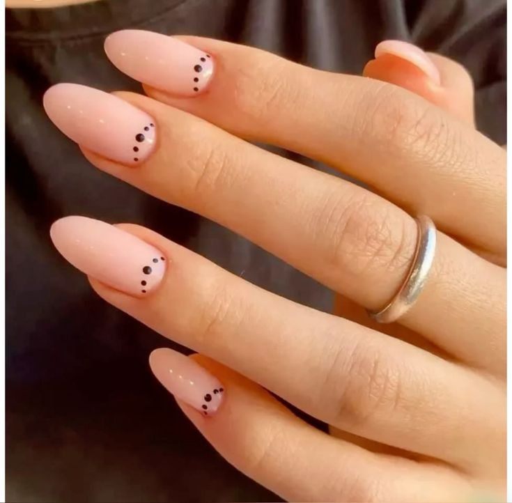 Unhas pintadas em tom nude, com pequenos pontos pretos, representando um design minimalista e elegante