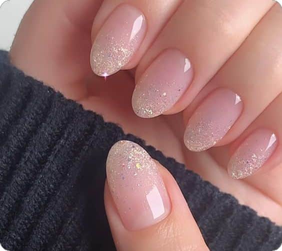 Unhas com esmalte em tom nude e um degradê de glitter dourado nas pontas, representando uma nail art minimalista e sofisticada.