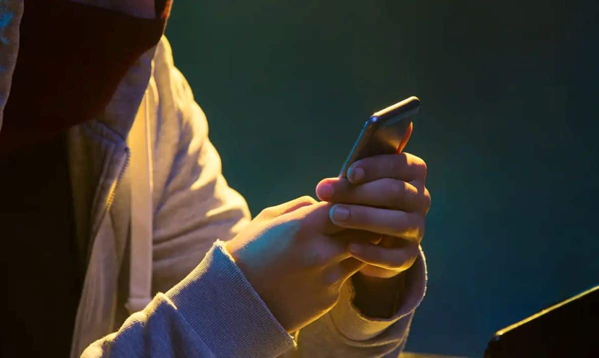 Pessoa com parte do rosto coberta por um capuz, segurando um smartphone com as duas mãos, em um ambiente de pouca luz. A imagem evoca a ideia de fraude digital, golpes e atividades suspeitas online.
