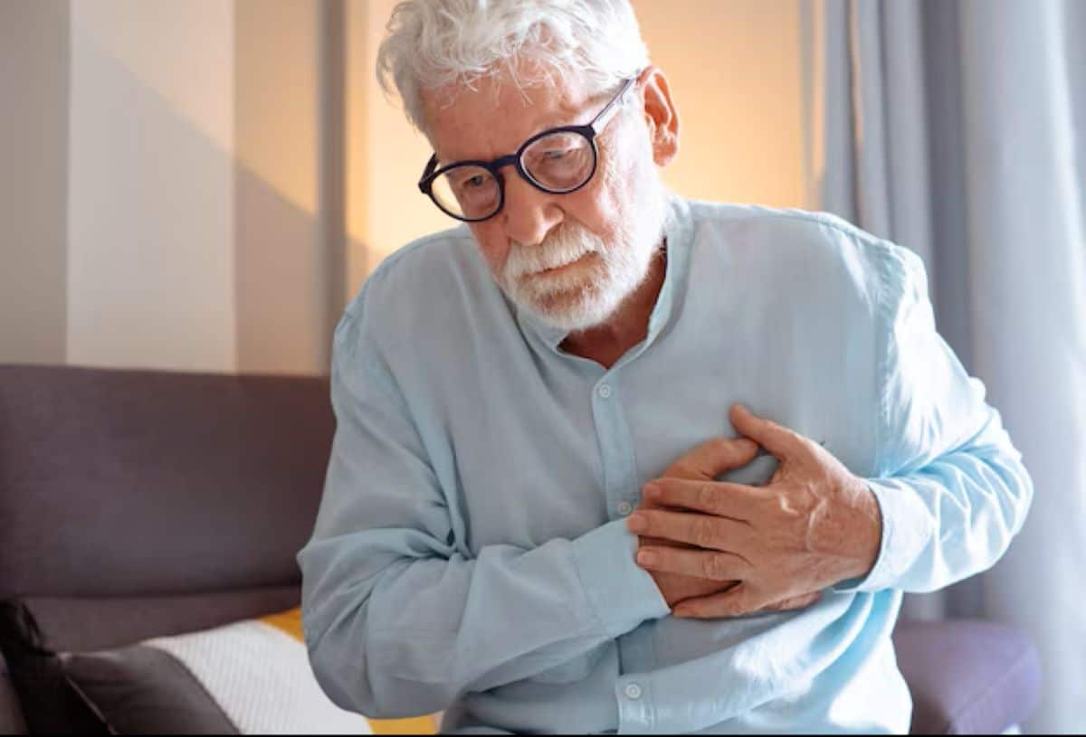 Homem idoso com óculos e camisa azul, expressando dor e segurando o peito, um sinal clássico de possível infarto ou angina.
