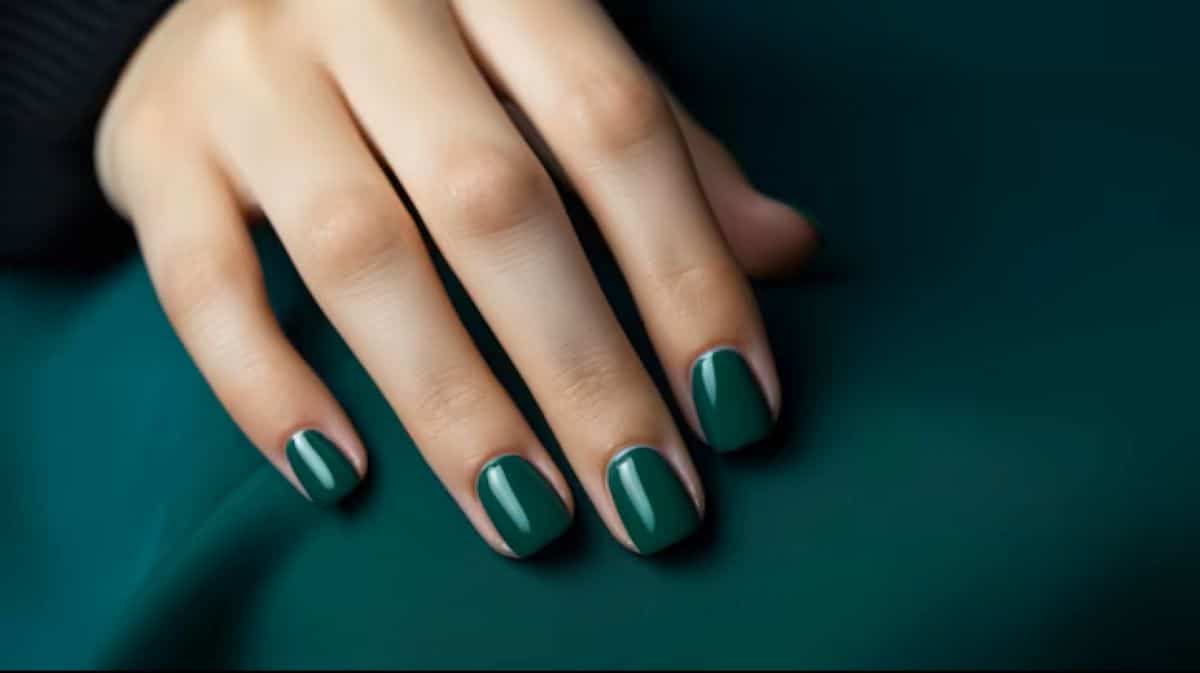 Mão feminina com unhas curtas pintadas de esmalte verde-escuro brilhante sobre tecido verde da mesma cor.