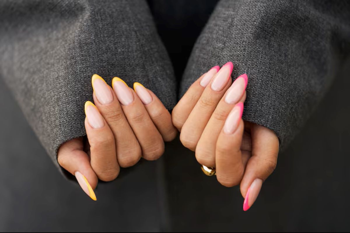 Mãos com unhas stiletto de ponta fina, decoradas com francesinhas coloridas em rosa neon e amarelo vibrante.