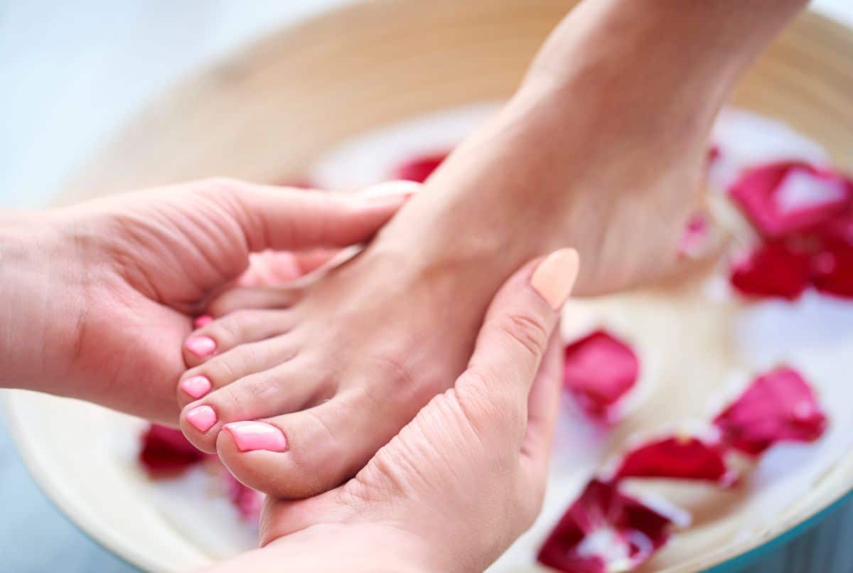 Pés femininos com unhas pintadas de rosa claro recebendo massagem e imersão em tigela de água com pétalas, ilustrando um tratamento de pedicure para alongar unhas.