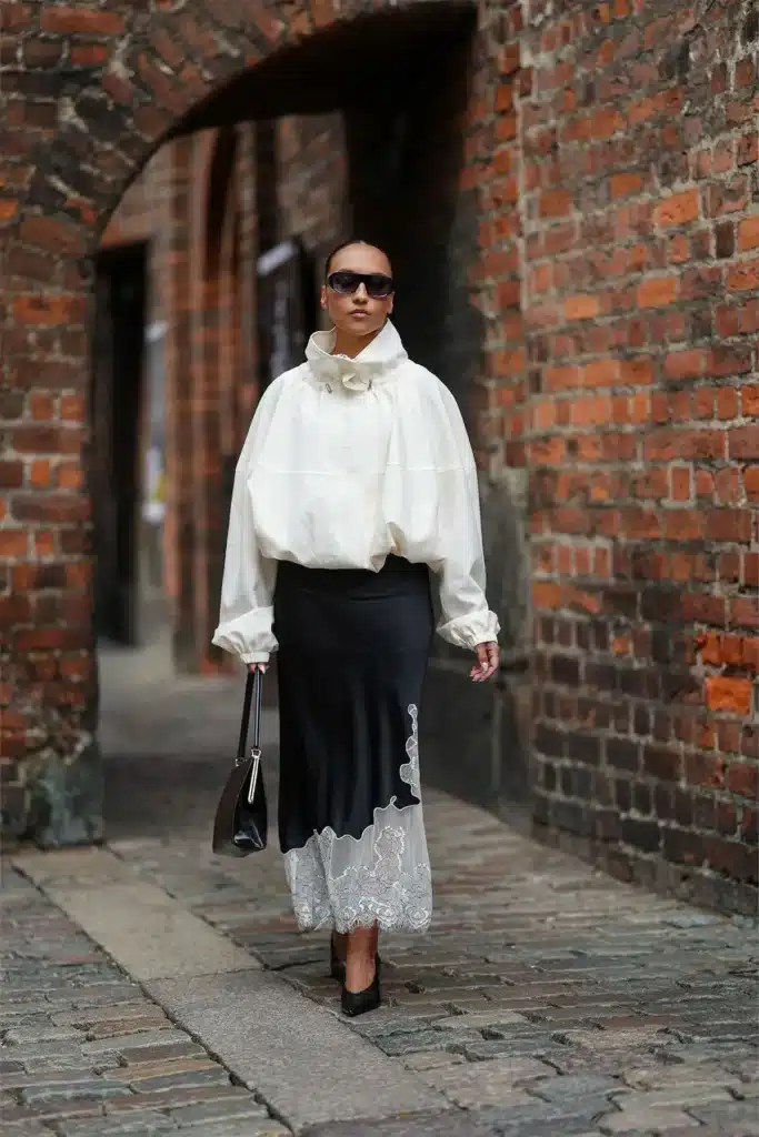 Look com blusa volumosa e saia de renda Mulher estilosa com blusa volumosa e saia com detalhes de renda, caminhando por uma rua de pedras.