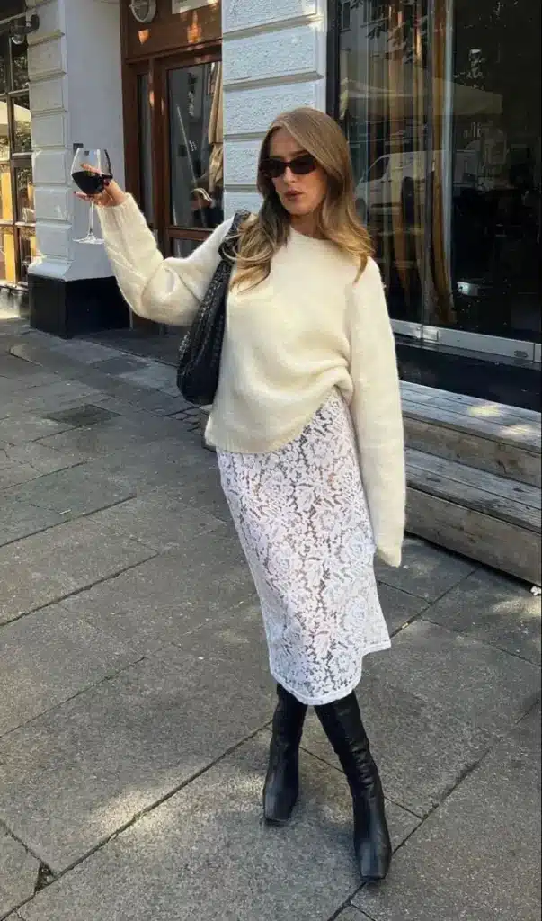Look de tricô e renda para um estilo moderno e elegante Mulher usando um conjunto de tricô oversized e saia de renda, segurando um copo de vinho tinto em um cenário urbano.