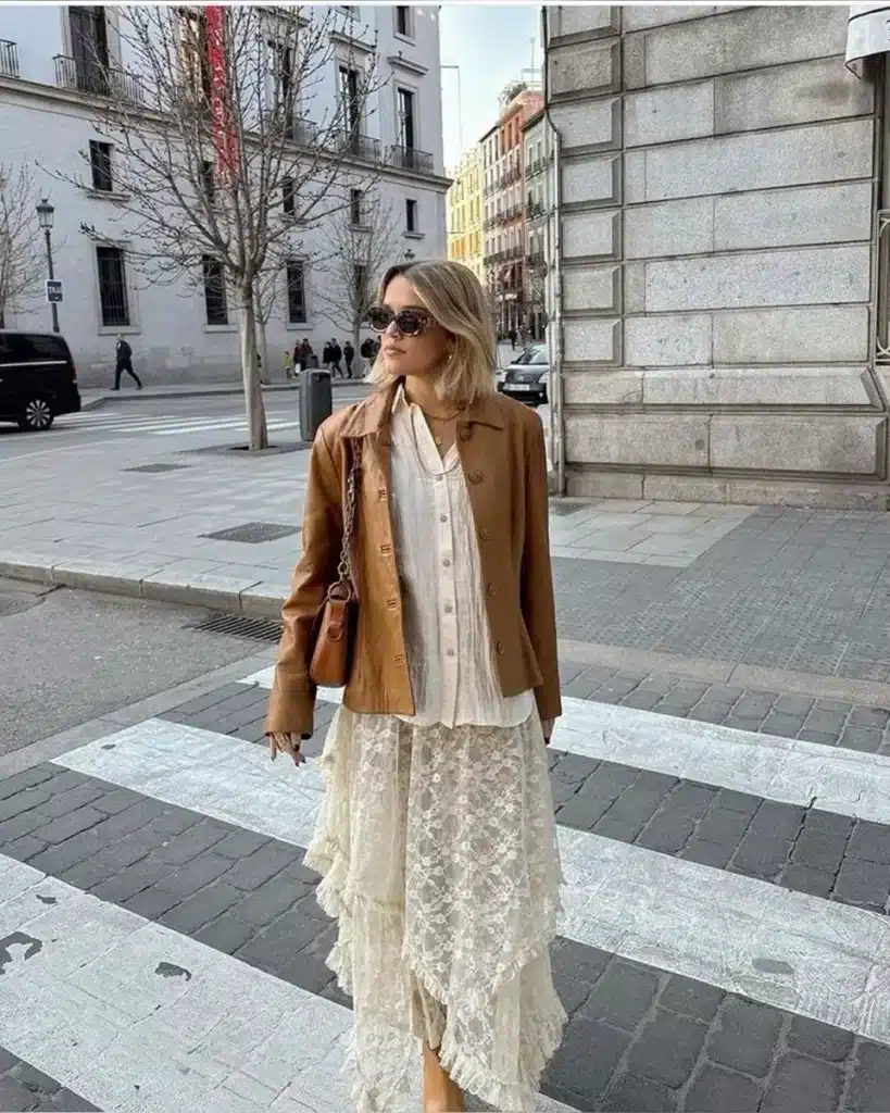 Estilo moderno: look com jaqueta de couro e saia assimétrica Mulher com jaqueta de couro e saia assimétrica de renda, cruzando a rua em uma cidade, com óculos escuros e bolsa de ombro.