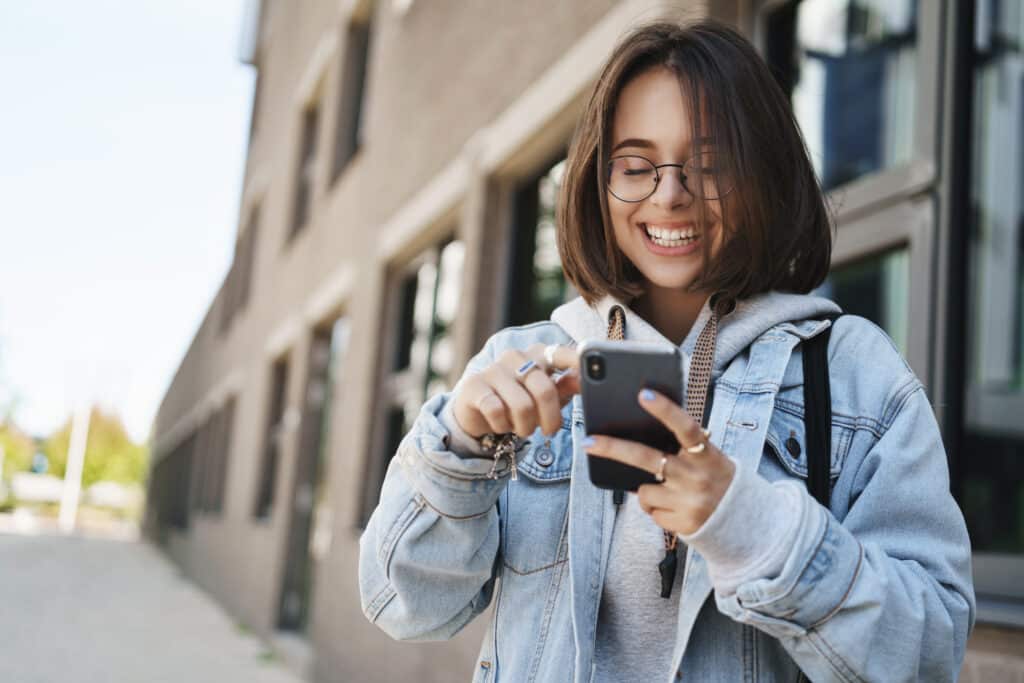 Mulher jovem com óculos e jaqueta jeans rindo enquanto olha para a tela do celular.