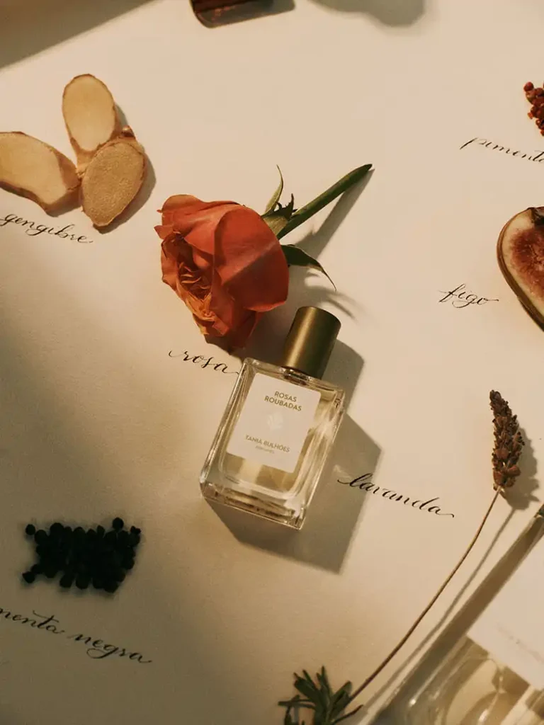 Frasco do perfume Rosas Roubadas, cercado por ingredientes como gengibre e pimenta, ilustrando as notas complexas das fragrâncias florais da Primavera.