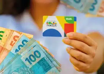 Imagem ilustrativa do Bolsa Família mostrando uma mão segurando o cartão do programa junto a notas de reais de diferentes valores, com destaque para as cédulas de 100, 50 e 20 reais.