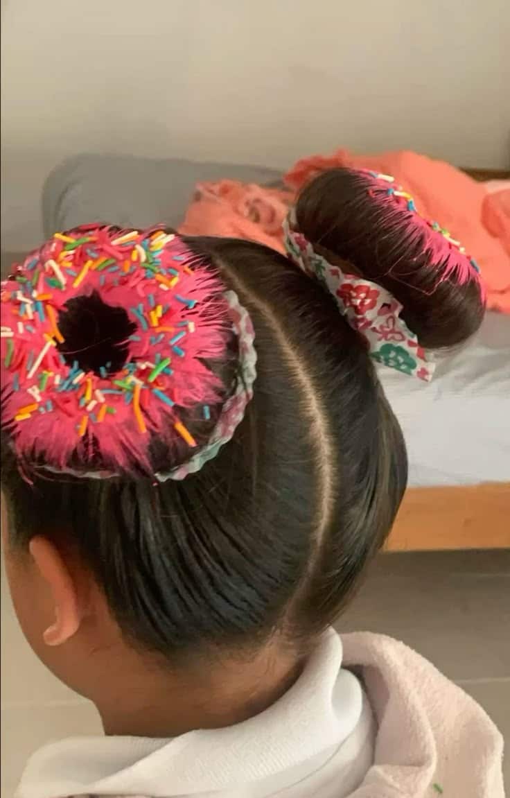 Menina com penteado de cabelo maluco de rosquinhas (donuts), usando dois coques decorados com spray rosa e granulado colorido para festa na escola.