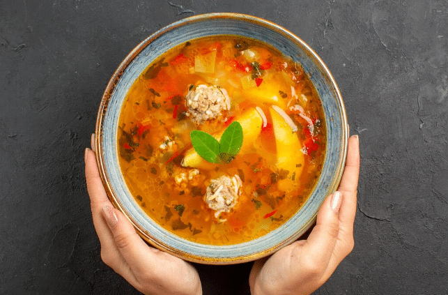 Tigela de sopa quente com almôndegas, legumes e ervas, segurada por duas mãos sobre fundo preto
