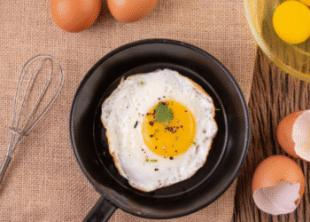 Ovo frito com gema mole na frigideira, rodeado por ovos crus, cascas, gemas separadas e utensílios. Ilustra a preparação de ovos fritos e o segredo do vinagre.