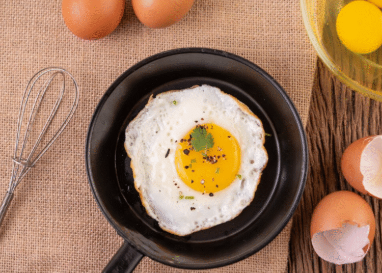 Ovo frito com gema mole na frigideira, rodeado por ovos crus, cascas, gemas separadas e utensílios. Ilustra a preparação de ovos fritos e o segredo do vinagre.