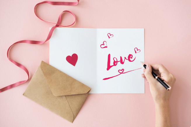 Mão escrevendo palavra Love em cartão branco com corações vermelhos e envelope kraft sobre fundo rosa