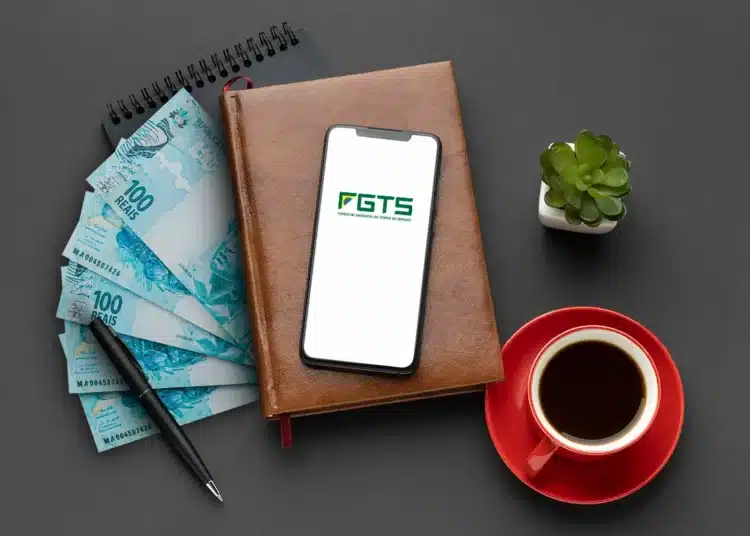Imagem mostrando notas de 100 reais, um smartphone com o logo do FGTS, um caderno, uma caneta e uma xícara de café. Tudo sobre um fundo cinza escuro.