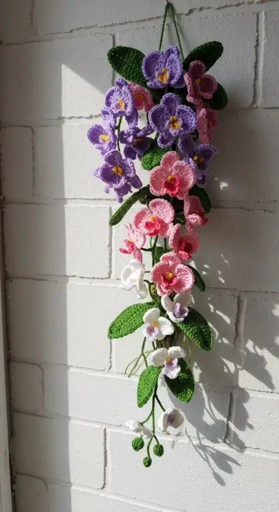 Orquídeas de crochê penduradas com flores em tons de rosa, roxo e branco, destacando folhas verdes detalhadas.
