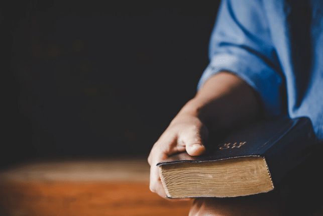 Mão segurando Bíblia preta em ambiente com fundo desfocado
