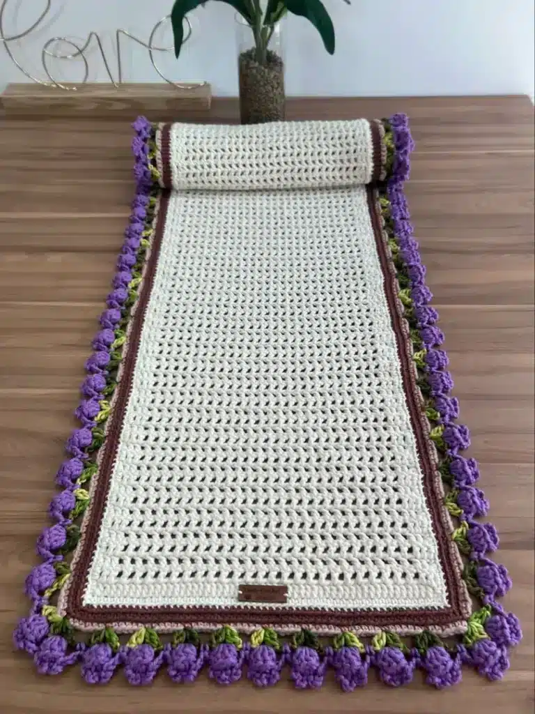 Trilho de crochê com flores nas pontas, com detalhes em roxo, verde e marrom, sobre uma superfície de madeira.