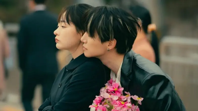 Cena de um momento emocional em Typhoon Family, com dois personagens próximos, com um jovem homem segurando flores enquanto está perto de uma mulher.