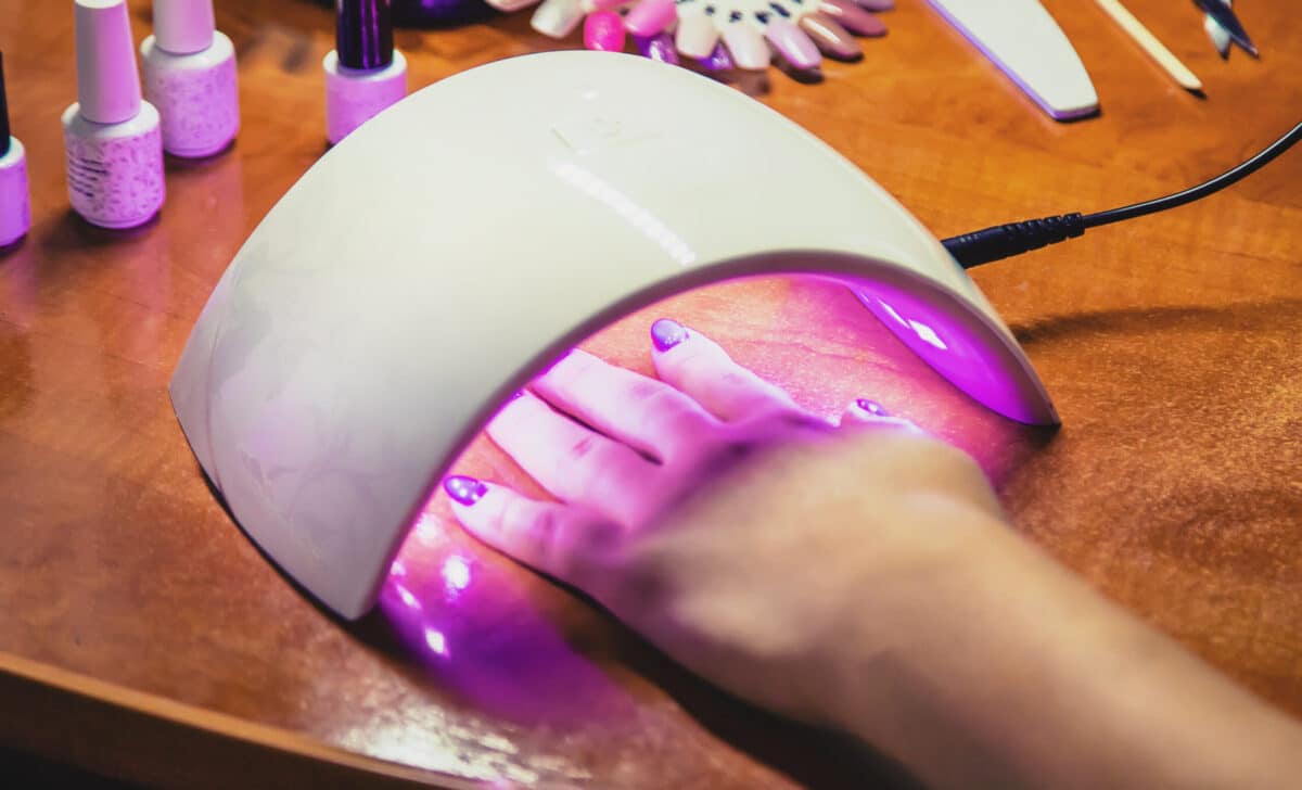 Mão de cliente em cabine UV/LED para secagem de unhas em gel, equipamento crucial no processo de polimerização que utiliza as substâncias TPO e DMPT.