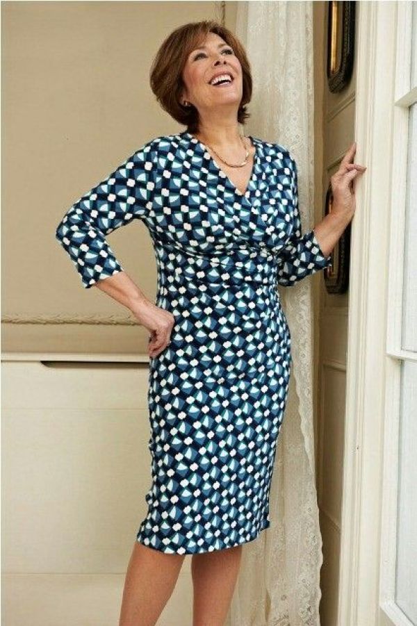 Mulher sorrindo em pose elegante usando um vestido transpassado ou envelope de estampa geométrica azul, verde e branca. A modelagem ajustável do vestido realça a cintura e alonga a silhueta.