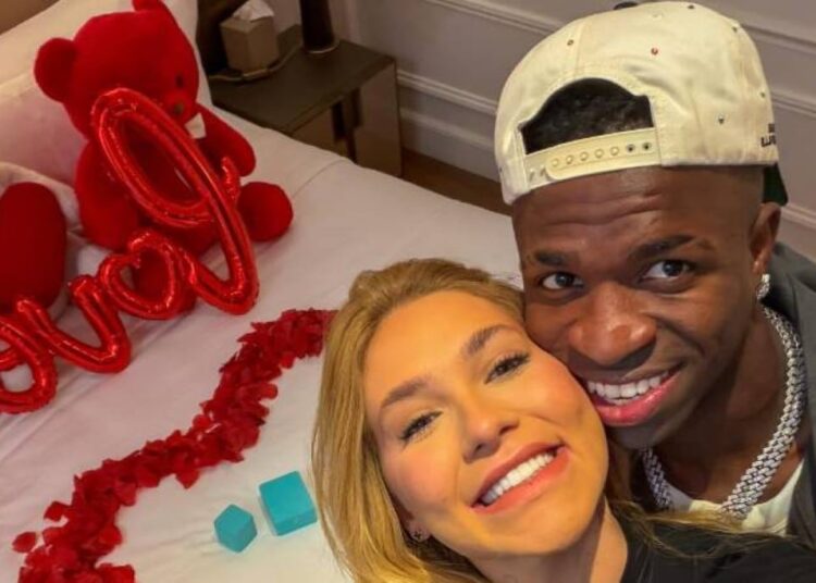 Homem e mulher sorrindo próximos à cama com decoração de ursinhos e balões vermelhos formando a palavra love