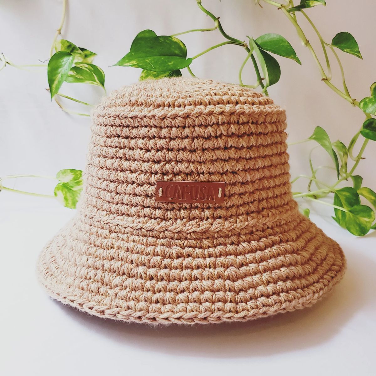 Chapéu bucket de crochê em tom bege neutro com etiqueta 'CAFUSA', exibindo textura artesanal e fundo com folhagem verde. Representa opções de acessórios feitos à mão disponíveis em 2025.