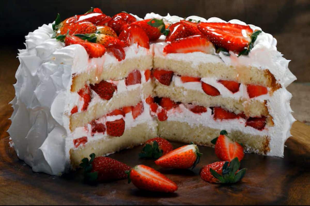Bolo inteiro de leite ninho com morango cortado ao meio, revelando múltiplas camadas de recheio cremoso e morangos frescos picados, decorado com chantilly e morangos espalhados ao redor em uma base de madeira escura.